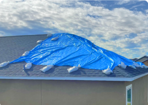 Homeowner placing a tarp over a leaking roof during heavy rain while emergency roof repair professionals prepare to fix storm damage and water leaks.